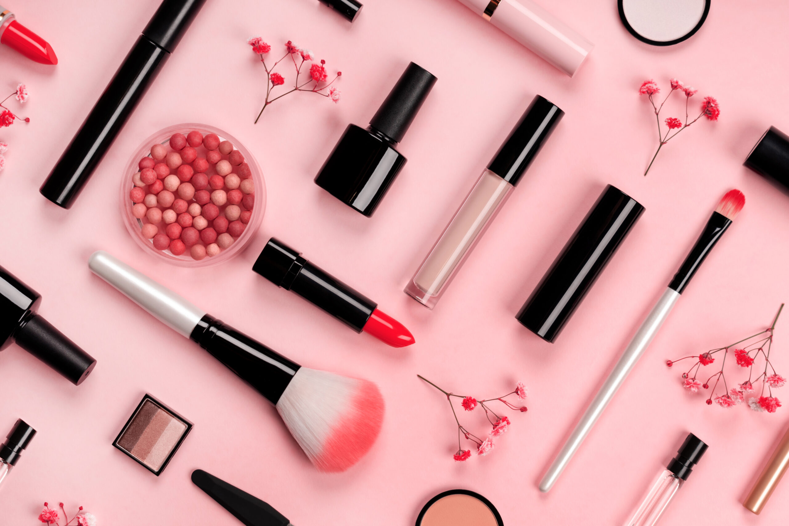 Various cosmetic accessories for makeup and manicure on trendy pastel pink background with red flowers. Blush, brush, eye shadow, mascara, perfume, lipstick, nail Polish. Skin care products.